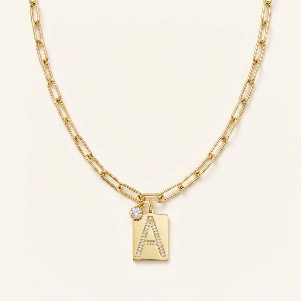 Pave initial pendant necklace hero — gold tag pendant pavé crystal letter A on paperclip chain, off-white neutral background matching reference