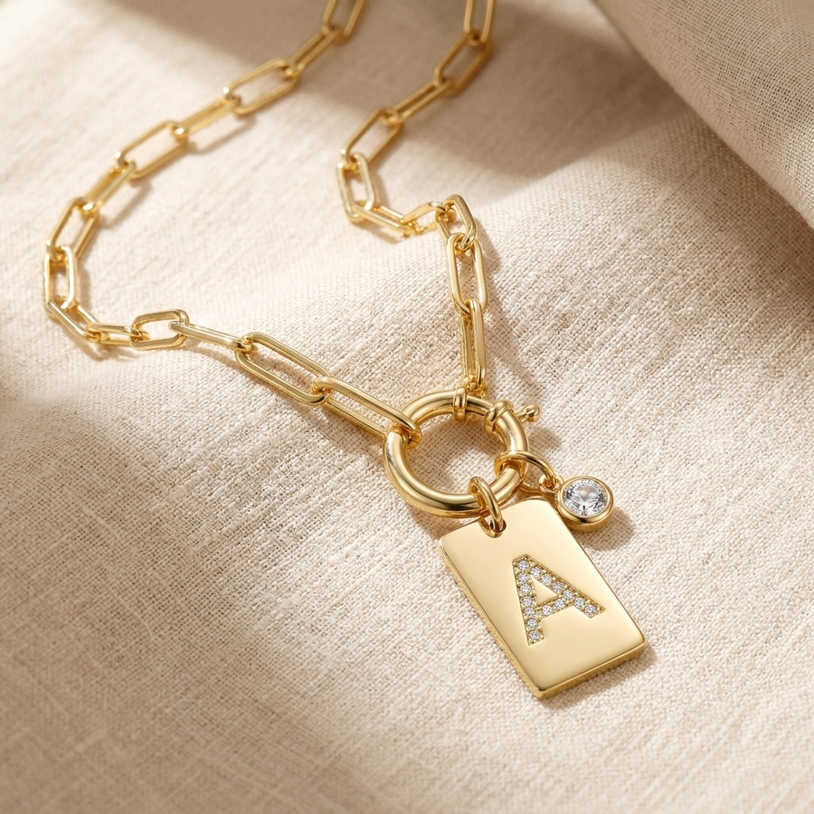 Pave initial pendant necklace on linen — gold pendant tag with pavé crystal letter A and paperclip chain coiled on linen fabric