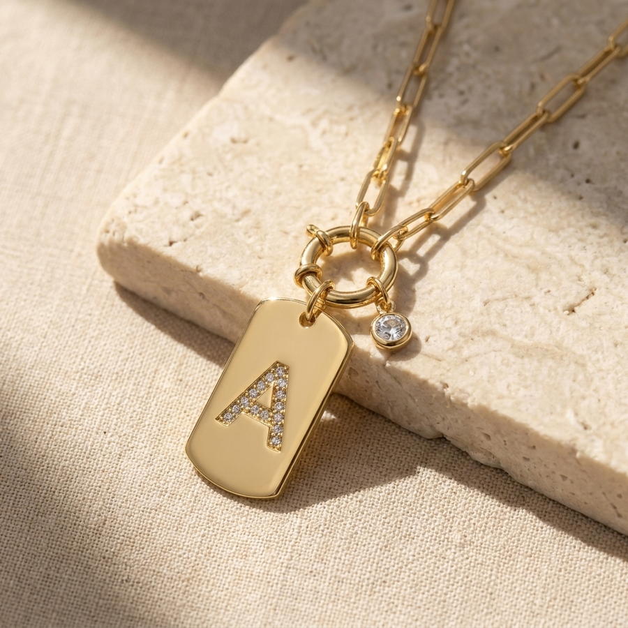 Pave initial pendant necklace on stone — gold pendant tag with pavé crystal letter A and paperclip chain draped on natural stone block