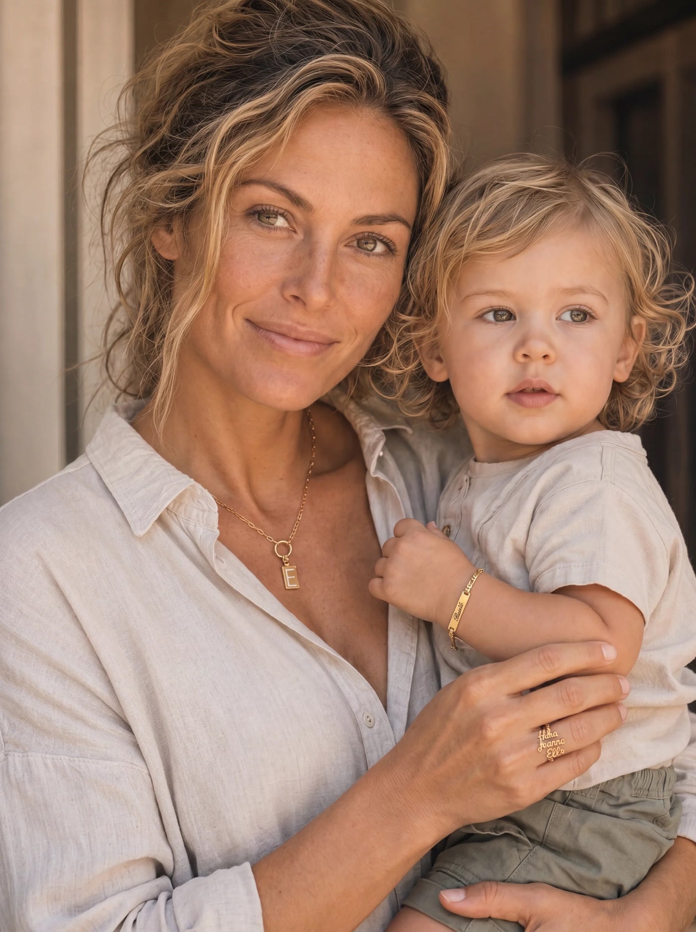 A mother in her late thirties holding her young blond toddler in warm window light, both in soft linen tones; she wears an Heirpearl gold initial necklace and a name-engraved ring, the child wears a delicate engraved Heirpearl bar bracelet.