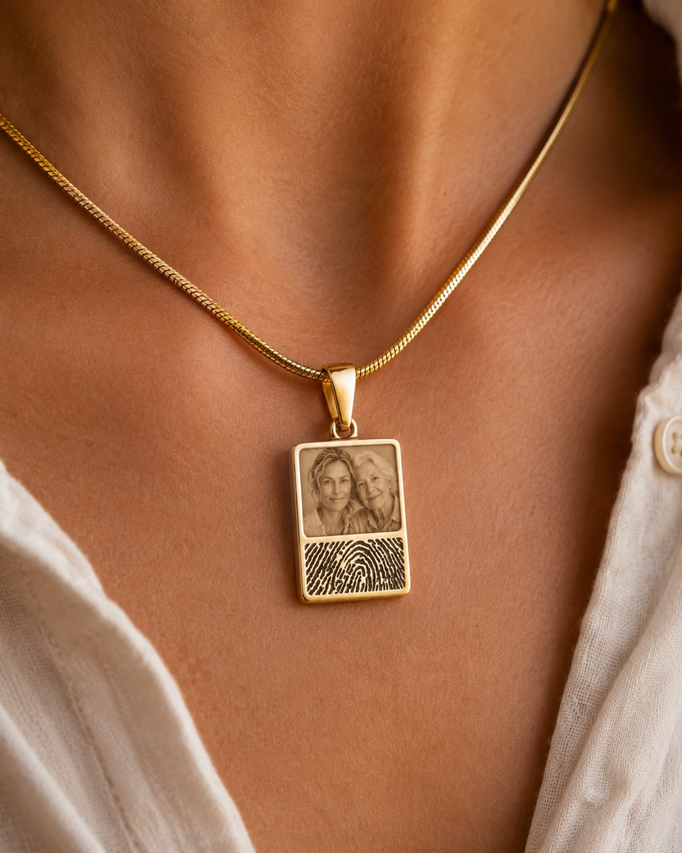 Macro close-up of a rectangular Heirpearl gold pendant resting on warm skin: the upper half holds a soft photograph of a loved one, the lower half a finely engraved fingerprint, on a slim gold snake chain.